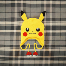 Load image into Gallery viewer, Pokemon Laplander Pikachu Beanie with Pikachu face, ears, and Poké Ball tassels on plaid background.
